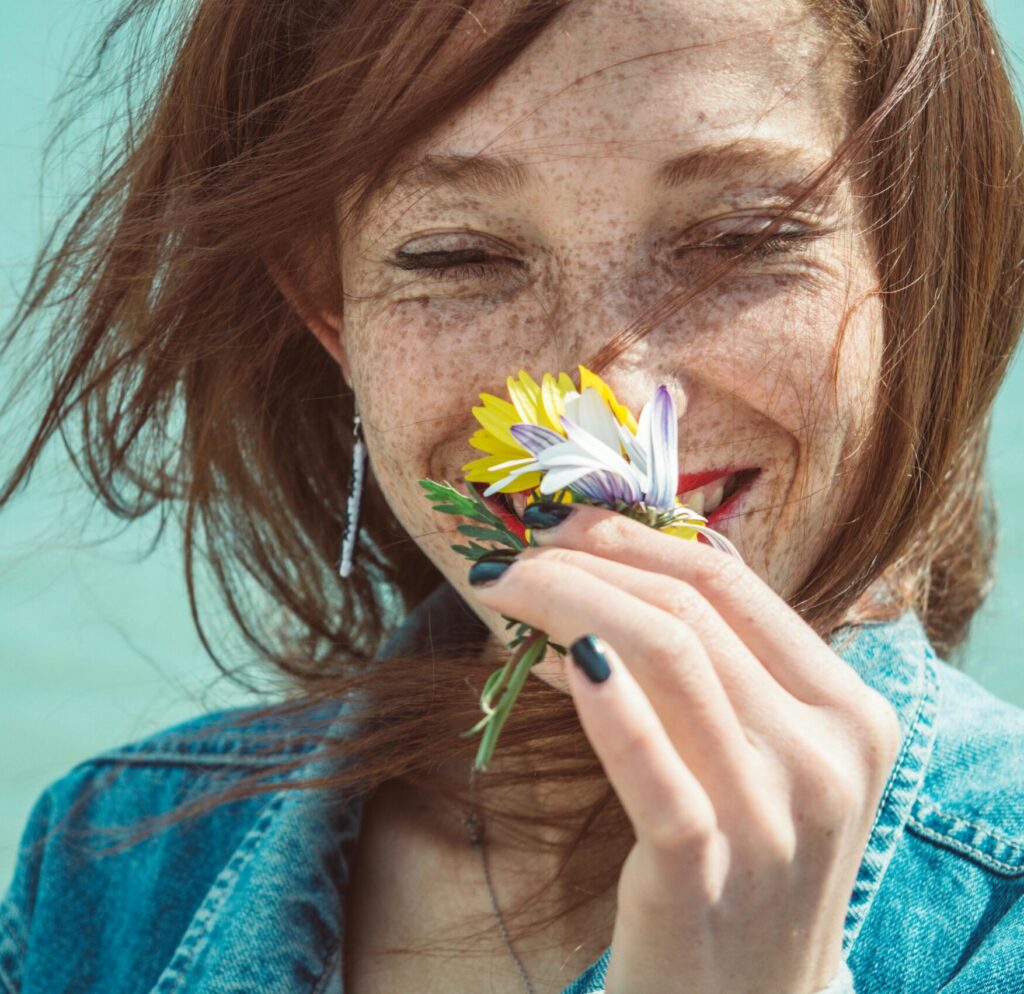 Smiling woman with freckles enjoying the aroma of flowers near a blue water background.