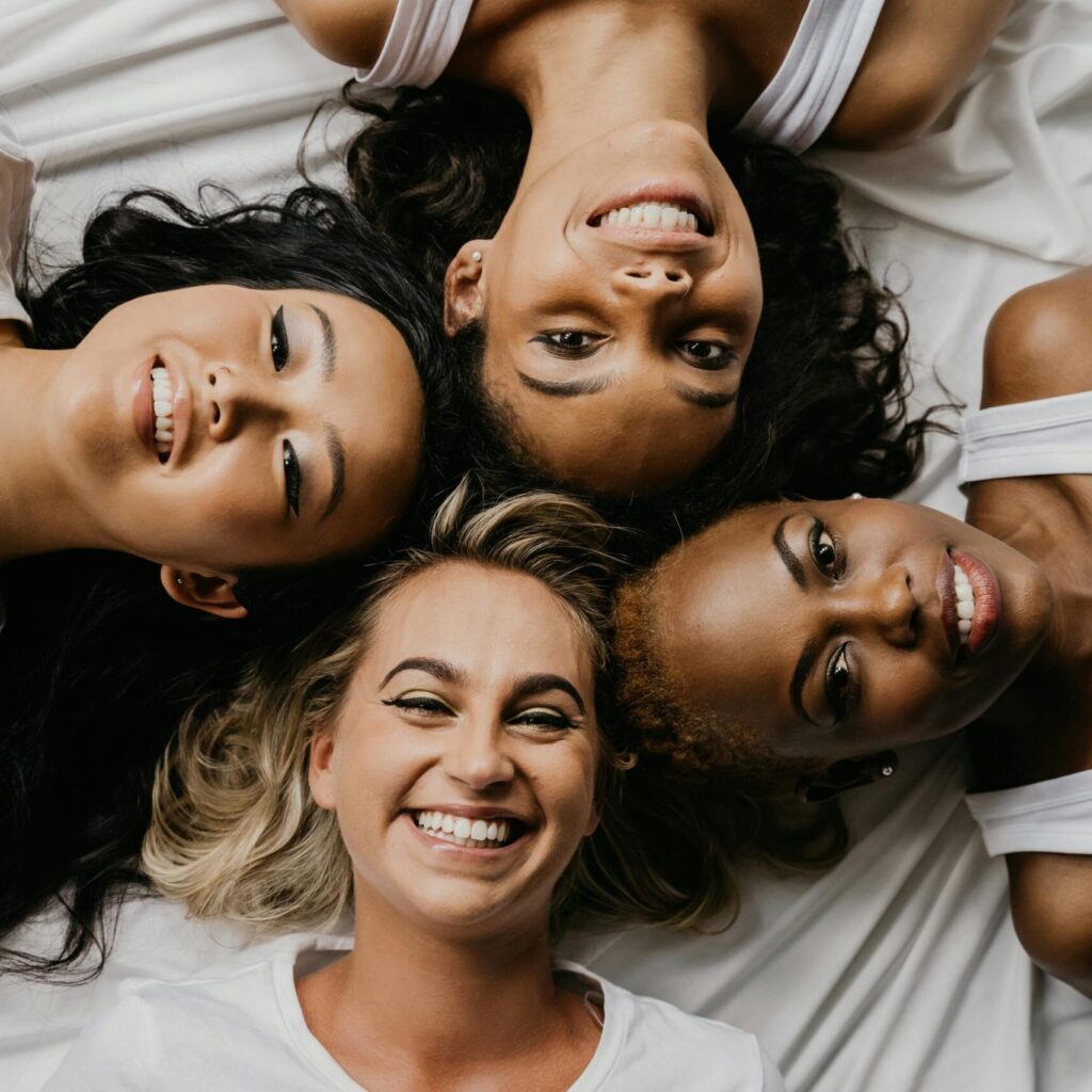 Four diverse women lying on a bed smiling at the camera, sharing a joyful moment.